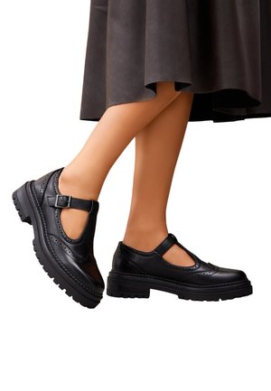 Jambes en collants couleur hâlée portant des chaussures Mary Jane en cuir noir avec des semelles épaisses et des brides à boucle, associées à une jupe foncée.