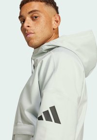 Ljusgrå hoodiejacka gjord av slätt tyg, med en svart logotyp på vänster ärm och dragkedja fram med en avslappnad passform.