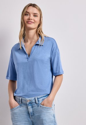 Giovane donna con capelli biondi che indossa una camicia celeste a maniche corte con colletto e jeans blu sbiaditi con le mani nelle tasche.