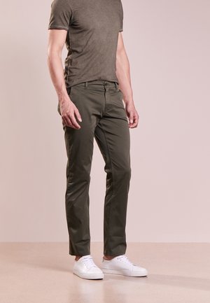 Hombre con pantalones ajustados color verde oliva, camiseta a juego verde oliva y zapatillas blancas, de pie contra un fondo claro y liso.