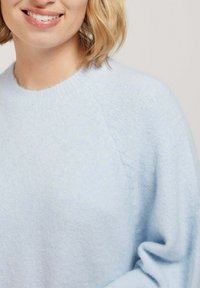Pull bleu clair avec un tricot doux et texturé. Il présente un col rond et des manches raglan, affichant une coupe décontractée et une variation subtile de couleur.