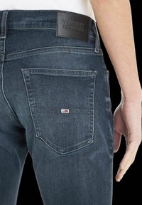 Jeans de mezclilla azul oscuro con un parche de cuero con la marca "TOMMY JEANS", bolsillo trasero con un pequeño detalle de logo y acentos de costura.