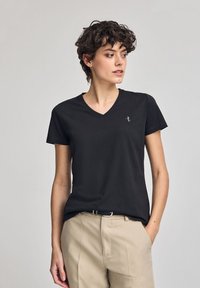 Svart bomullströja med v-ringad hals och korta ärmar; har en liten broderad design på vänster bröst. Del av en ensemble med beige byxor.