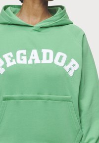 Sudadera verde de tejido suave y texturizado. Presenta la palabra "PEGADOR" en letras blancas en el pecho y un bolsillo tipo canguro en la parte frontal.