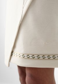 Nike krilo v kremni barvi z teksturirano tkanino, ki ima okrasni tkani rob v zeleni in beli barvi. Opazna je subtilna blagovna znamka.