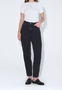 Femme portant un haut blanc à manches courtes, un jean noir taille haute, des chaussures noires sans lacets et un collier pendentif, debout devant un fond blanc.