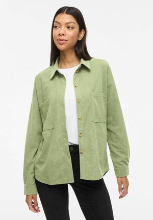 Vrouw die een lichtgroen corduroy overhemd met knopen draagt over een wit shirt en een zwarte broek, staand met een ontspannen houding tegen een witte achtergrond.