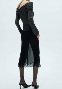 Off-shoulder donkergrijze gebreide top gecombineerd met een zwarte rok met kantaccenten en een doorzichtige paneel. Zwarte gekpunt hakschoenen maken de look compleet.
