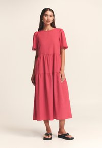 Abito maxi fucsia a balze con maniche a palloncino corte e scollatura rotonda, realizzato in tessuto leggero. Abbinato a sandali neri.