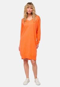 Robe orange à manches longues avec un décolleté en V et une coupe décontractée. Fabriquée en matériau doux, elle présente des poignets et un ourlet côtelés. Associée à des baskets blanches.