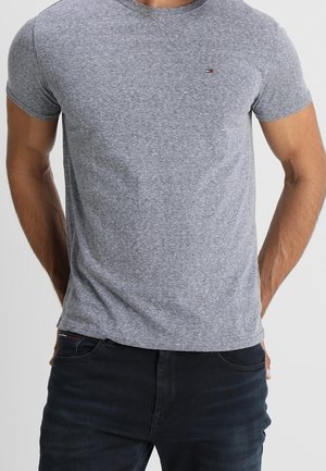 Mand iført en tætsiddende grå kortærmet t-shirt med lille logo på brystet og mørke jeans med hænderne i baglommerne.