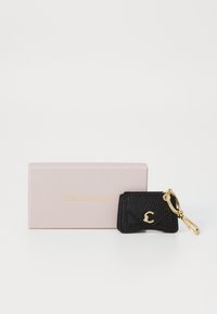 Porte-clé en cuir noir texturé avec un accent logo doré, attaché à un anneau de clé doré, positionné à côté d'une boîte cadeau rose avec un texte doré.