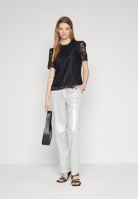 Zwart kanten top met korte pofmouwen, golvende halslijn en zilveren wijde broek met een gestructureerde afwerking. Zwarte handtas en sandalen met bandjes.