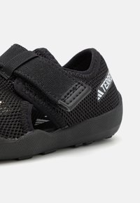 Zapato de malla negra con una correa de velcro, suela de goma texturizada y detalle de logo. Diseño ligero adecuado para diversas actividades.