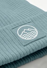 Knit beanie in light blue with ribbed texture, featuring a rectangular fabric patch with a white mountain and wave logo.