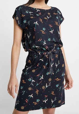 Kvinde iført en mørkeblå kjole med korte ærmer og blomsterprint, bundet med en flettet snor i taljen.