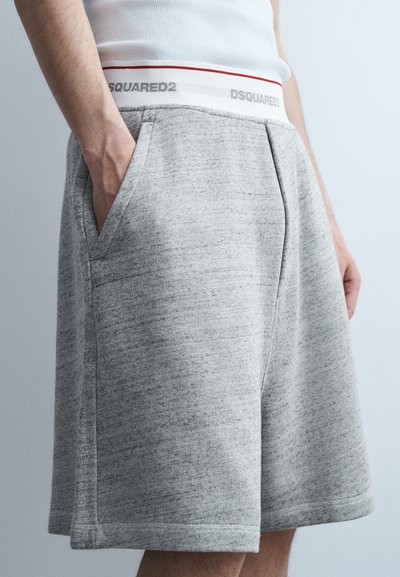 Homme portant un short décontracté gris avec des poches latérales et une ceinture blanche étiquetée "DSQUARED2", les mains dans les poches.