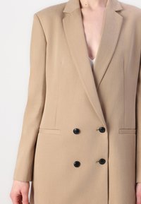 Beige dobbeltspent blazer med notched lapels, svarte knapper og sidelommer. Glatt tekstur, skreddersydd passform og moderne design.