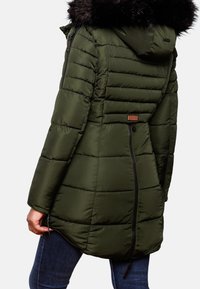 Femme portant un manteau d'hiver matelassé vert olive avec capuche bordée de fourrure et fermeture éclair au dos, associée à un jean bleu foncé, vue de dos.