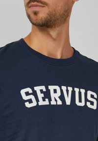 Granatowa koszulka z bawełny z wyraźnym białym napisem "SERVUS" na przodzie, okrągłym dekoltem i krótkimi rękawami.