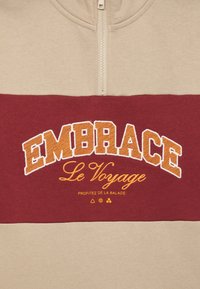 Beige og maroon sweatshirt med halvlukning, forsynet med broderet tekst "EMBRACE Le Voyage" i orange og gul. Glat tekstur.