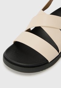 Beige kožené sandály s třemi širokými křížovými popruhy, černou texturovanou stélkou a plochou černou gumovou podrážkou. Jednoduchý design pro běžné nošení.