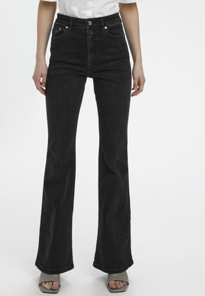 Persoon draagt zwarte, hoog getailleerde wijde jeans, wit overhemd ingestopt, en grijze sandalen met open tenen en hak, staande tegen een witte achtergrond.