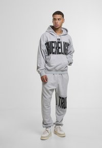 Súprava šedej mikiny a teplákov vyrobená z mäkkého materiálu. Mikina má veľký čierny text; tepláky majú zodpovedajúce čierne textové akcenty. Neformálny dizajn.