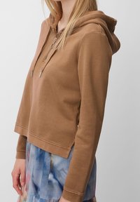 Hnědá sv hooded mikina vyrobená z měkké látky, s kapsou na zip a šňůrkou na dotažení. Střih je mírně zkrácený, stylizovaný s vrstvenou sukní.