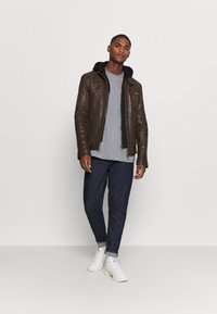 Giubbotto di pelle marrone con cappuccio nero, t-shirt grigia sotto, abbinato a jeans scuri e sneakers bianche. Tasche con zip sul giubbotto.