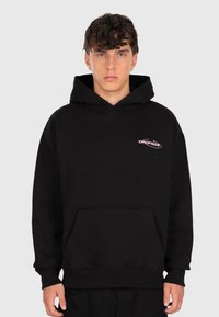 Jeune homme portant un sweat à capuche noir avec une poche avant et un petit logo rose "DROPSIZE" sur la poitrine gauche, devant un fond uni.