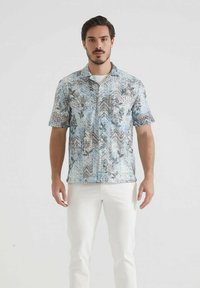 Homme se tenant contre un fond blanc portant une chemise à motifs bleue et grise à manches courtes avec un pantalon blanc.
