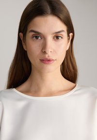 Satin white top with a scoop neckline, soft draping, and smooth texture. Model displays a natural makeup look with loose, straight hair.