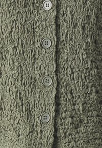 Tejido de color verde oliva con textura y superficie arrugada, que presenta cinco botones grises en la parte delantera para el cierre.