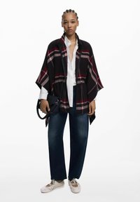 Schwarzes und rotes karierter Poncho mit lässiger Passform, getragen über einem weißen Hemd und kombiniert mit dunkler Jeans. Es hat eine große Vorderöffnung und Armlöcher.