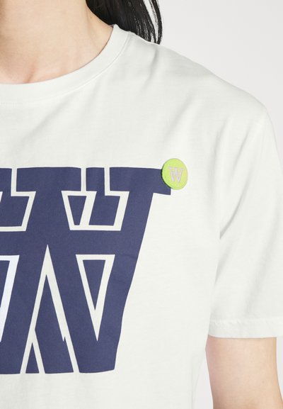 Balta medvilninė T-marškiniai su dideliu tamsiai mėlynu grafiniu dizainu, kuris turi geometrinę struktūrą. Pridėtas žalias mygtukas su "W" logotipo akcentu.