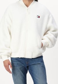 Een persoon in een wit, fluffy ritsjack met ribbels aan de manchetten en een klein Tommy Jeans-logo op de borst, gecombineerd met blauwe spijkerbroek.