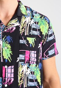 Camiseta negra de manga corta con patrones coloridos de palmeras y ventanas en rosa, verde y azul. Cuenta con un bolsillo en el pecho y cierre de botones.