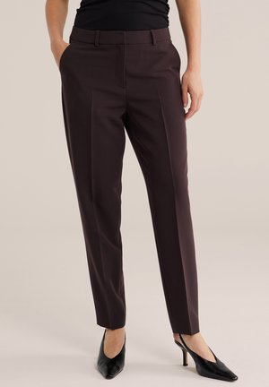 Kvinde iført mørke skræddersyede bukser med frontfold, sorte spidse slingback-hæle og sort top, hænderne afslappede ned langs siderne.