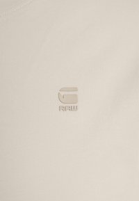 Petit logo G-RAW embossé beige sur fond de tissu beige clair.