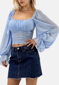 Hellblaues durchsichtiges Oberteil mit aufgeblähten Ärmeln und geraffter Textur am Oberkörper. Kombiniert mit einem dunklen Jeans-Minirock, ausgefranstem Saum und taillierter Passform.