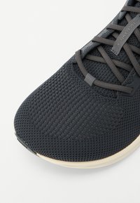 Chaussure de sport noire avec un dessus en mesh texturé, des lacets contrastants et une semelle légère. Caractérisée par un design épuré et un branding minimal.