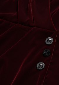 Burgundy samettimekko, jossa on laskoksia, ja kolme koristeellista mustaa nappia, joilla on teksturoitu muotoilu: kaksi pyöreää ja yksi pyöreä.