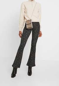 Svarta utsvängda byxor med ett vitt abstrakt mönster, matchat med en ljusbeige fleece zip-up tröja och en liten metallisk axelväska.