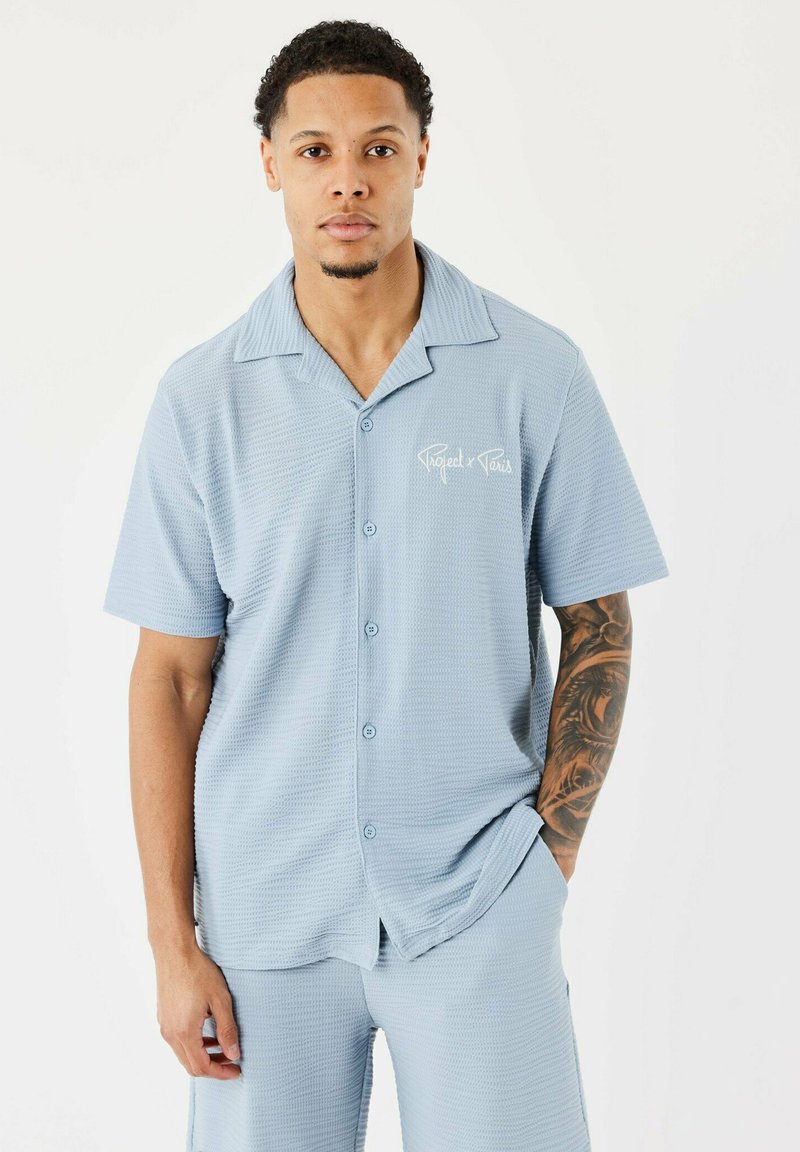 Chemise courte à manches courtes bleu clair avec un motif texturé, dotée d'une fermeture à boutons et d'un logo discret sur la poitrine. Associée à un short assorti.
