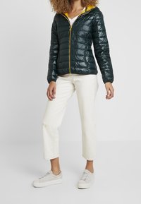 Mörkgrön puffjacka med en gul huva, dragkedjestängning och elastiska ärmmuddar; tillsammans med vita cropped byxor och ljusgrå sneakers.