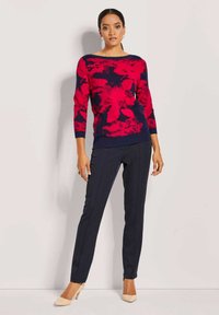 Rode bloempatronde gebreide sweater met een marineblauwe achtergrond, bootnek, en marineblauwe ribboorden, gecombineerd met op maat gemaakte donkere broeken en beige hakken.