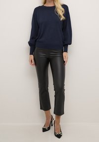 Marineblauer Pullover mit lockerer Passform und strukturiertem Finish, kombiniert mit eleganten, schwarzen, cropped Kunstlederhosen und schwarzen, schmalen Schuhen mit goldenen Akzenten.