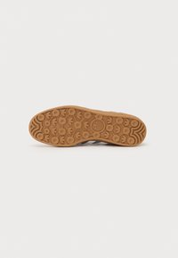 Semelle d’une chaussure Adidas marron avec des motifs circulaires et en forme de trèfle, avec le logo Adidas au centre sur un fond uni.