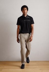 Tricou negru cu mânecă scurtă și text grafic, pantaloni bej și pantofi negri. Include, de asemenea, o eșarfă de gât cu model alb și negru.